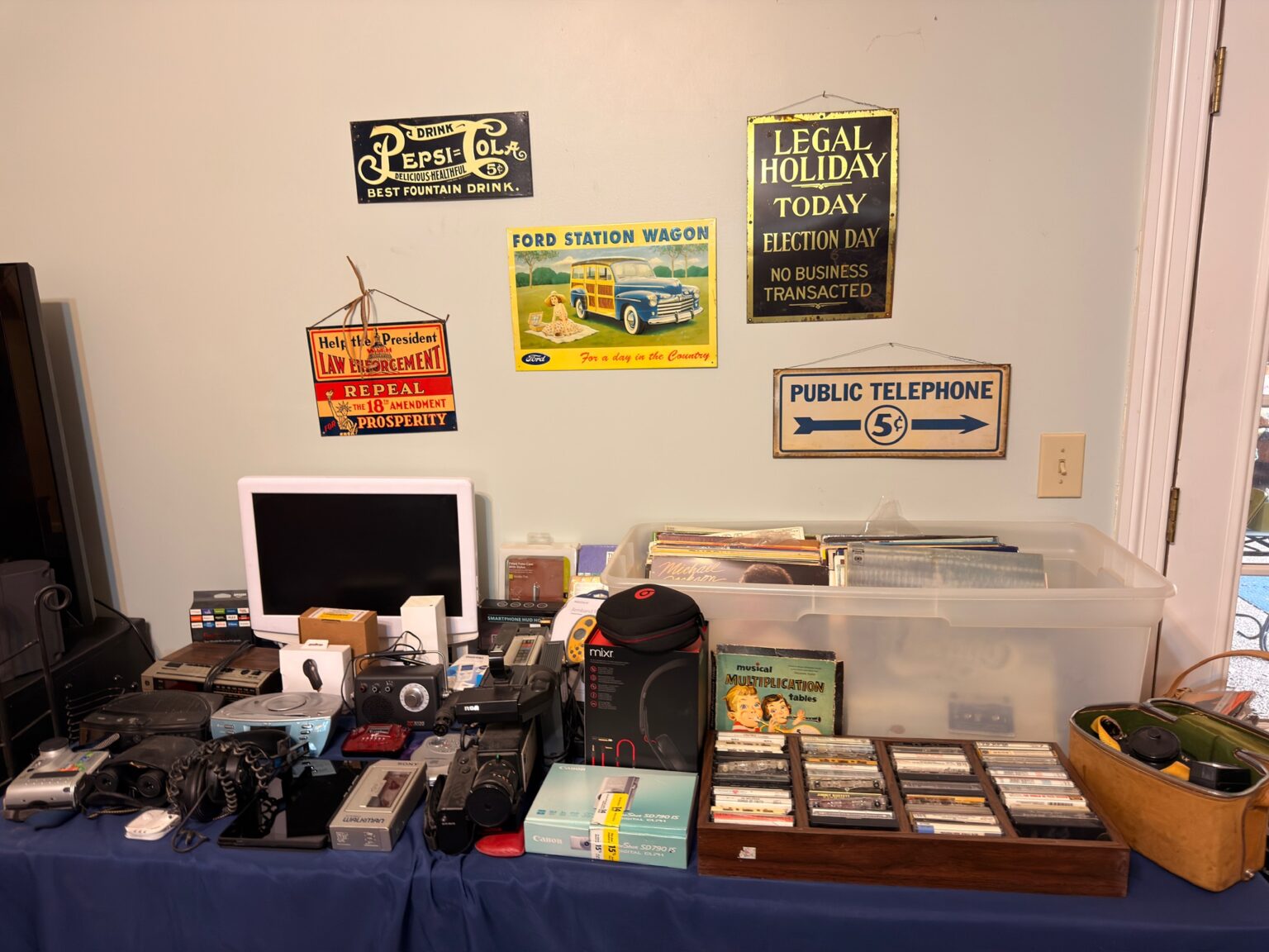 Vintage advertising signs above a table of records, cassette tapes, cameras, and electronics at an Estate Greats sale
